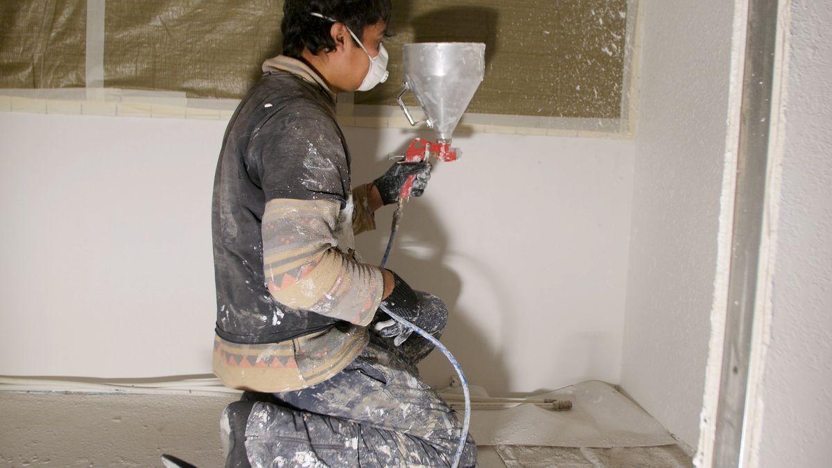 Worker painting wall with spray gun in white color. Man painting a grey wall, renovating exterior walls of new house. Open container with a thick white paint. Spray paint work